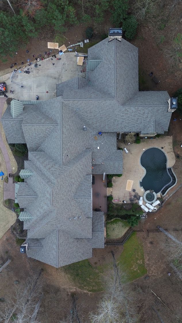 Aerial view of a large residential home with a complex shingled roof, a swimming pool, and a surrounding wooded lot.