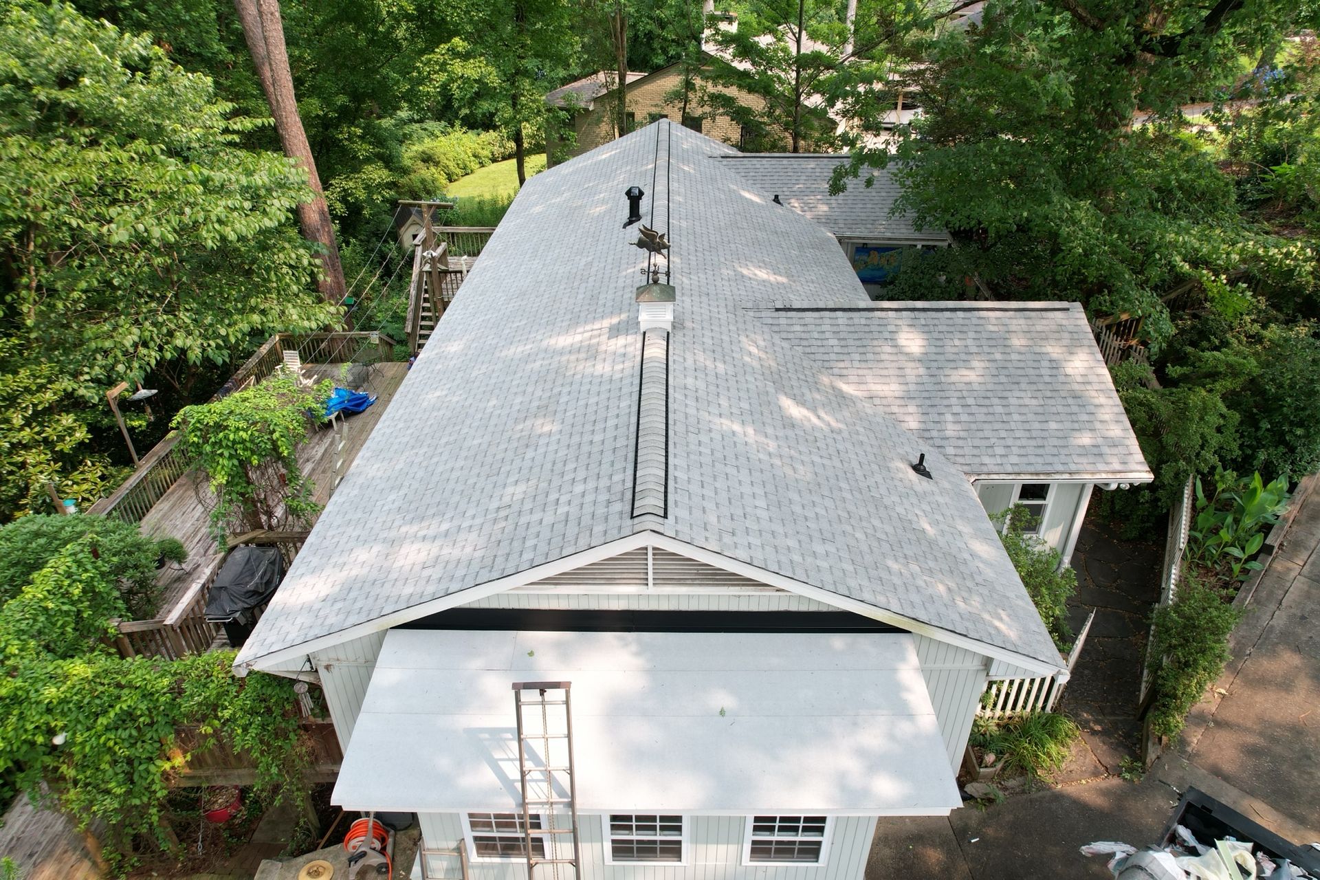 Aerial view of a gray shingled roof with a metal ventilation ridge vent, set in a lush, wooded residential area.