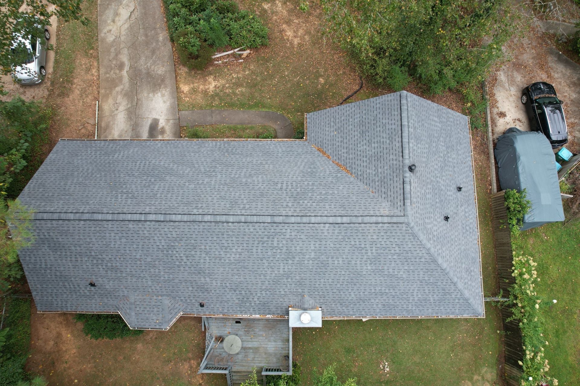 Aerial view of an L-shaped house with a grey shingled roof, surrounded by a yard, driveway, and parked vehicles.