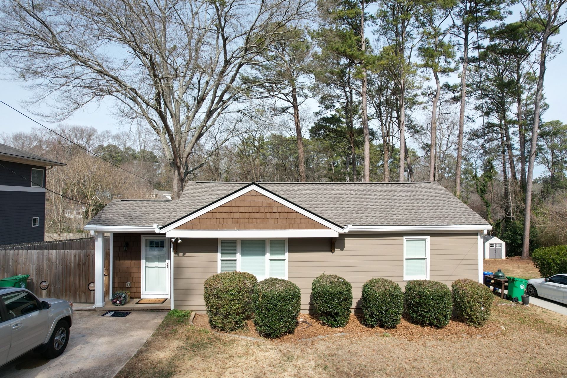 A single-story, tan-sided house with a brown triangular gable and a white door, situated in a treed residential area.