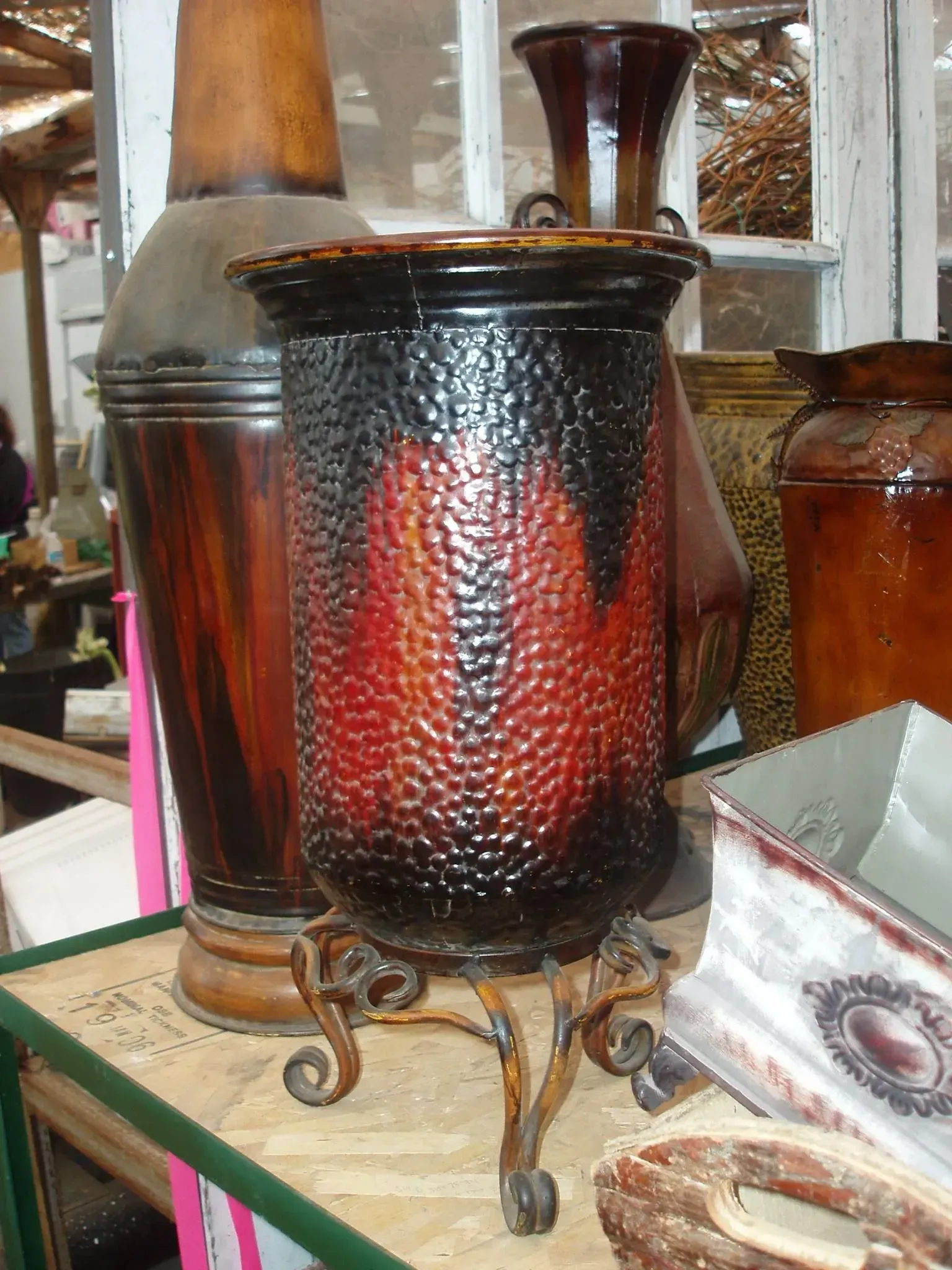 Red and black textured vase with ornate metal stand, various vases in background.