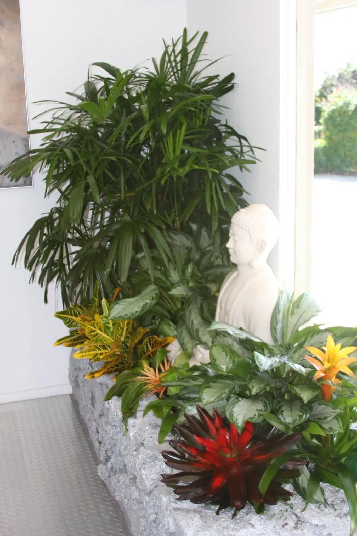 Statue of Buddha in planter with various green and orange plants, by a window.