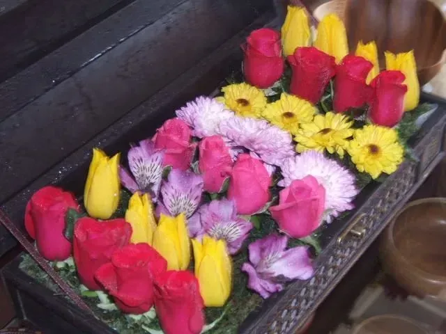 Assorted colorful flowers arranged inside a dark rectangular container, including red roses and yellow tulips.