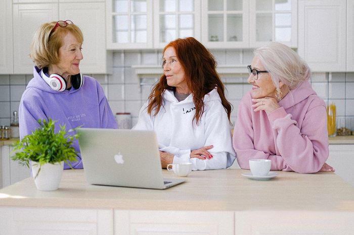 Three women smiling, gathered around a laptop on a kitchen counter. One wears headphones.
