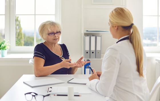 Woman talking to a doctor at a desk, light-filled office. The doctor has a stethoscope.