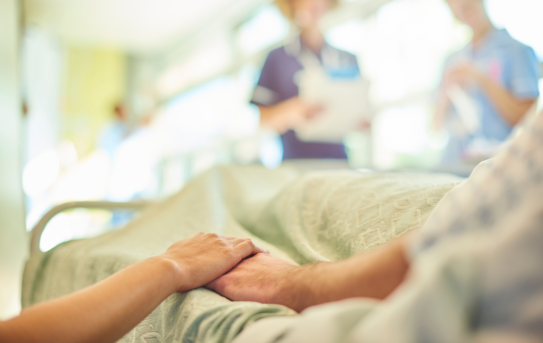 Hands clasped on a hospital bed, nurses in background.