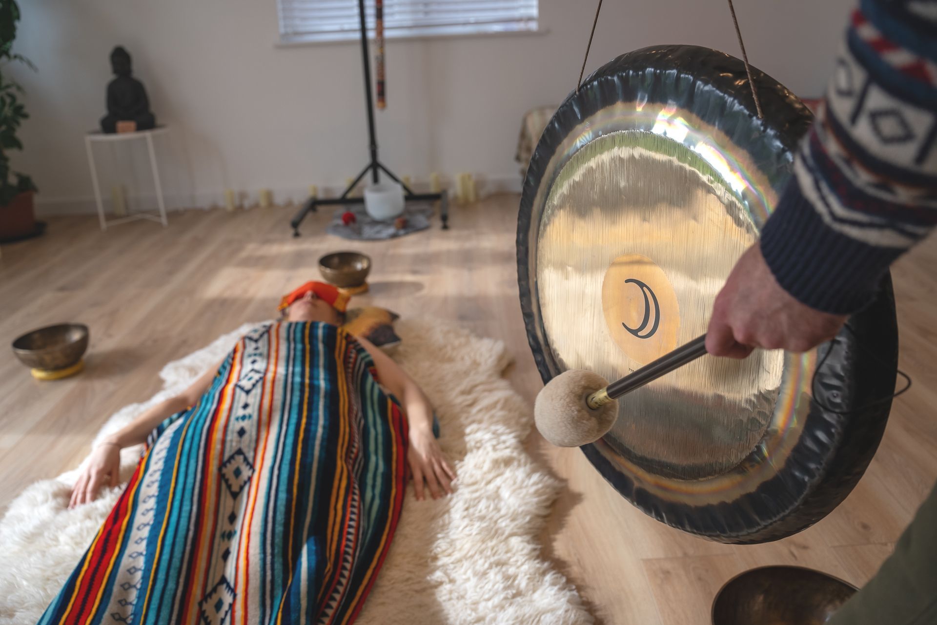 Person lying on rug, covered by blanket, near large gong being struck with mallet in light room.