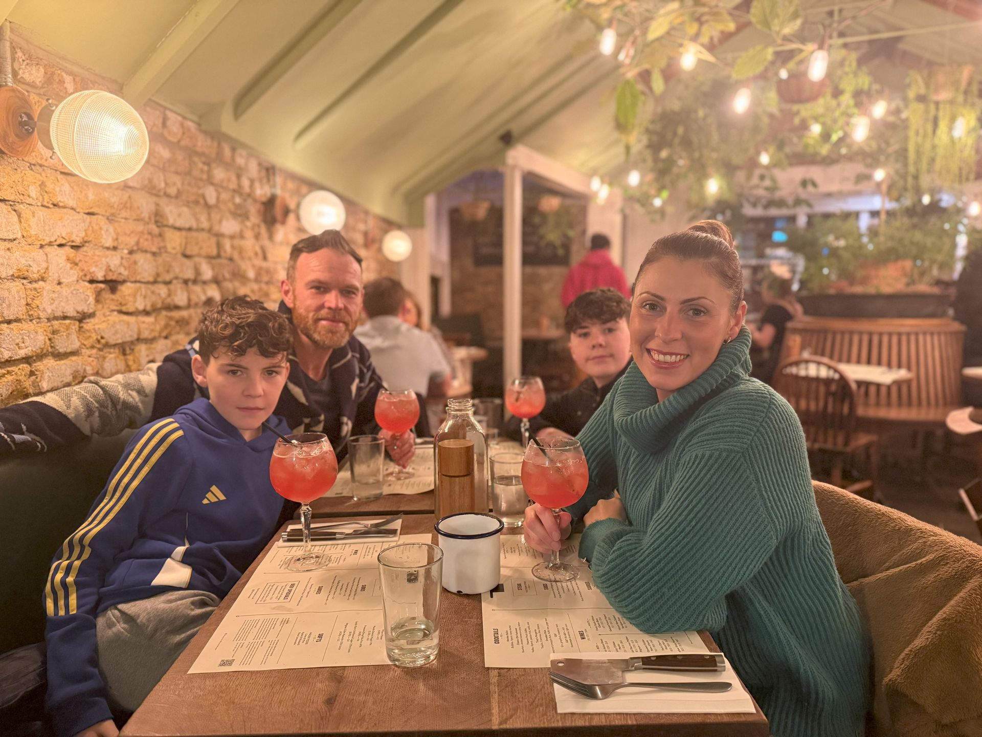 Family seated at a restaurant table, holding drinks. Interior with brick wall, overhead lights, and greenery.