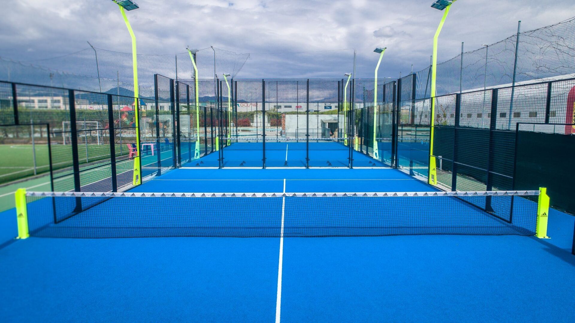 Campi da padel blu con reti e pareti di vetro, sotto un cielo nuvoloso.