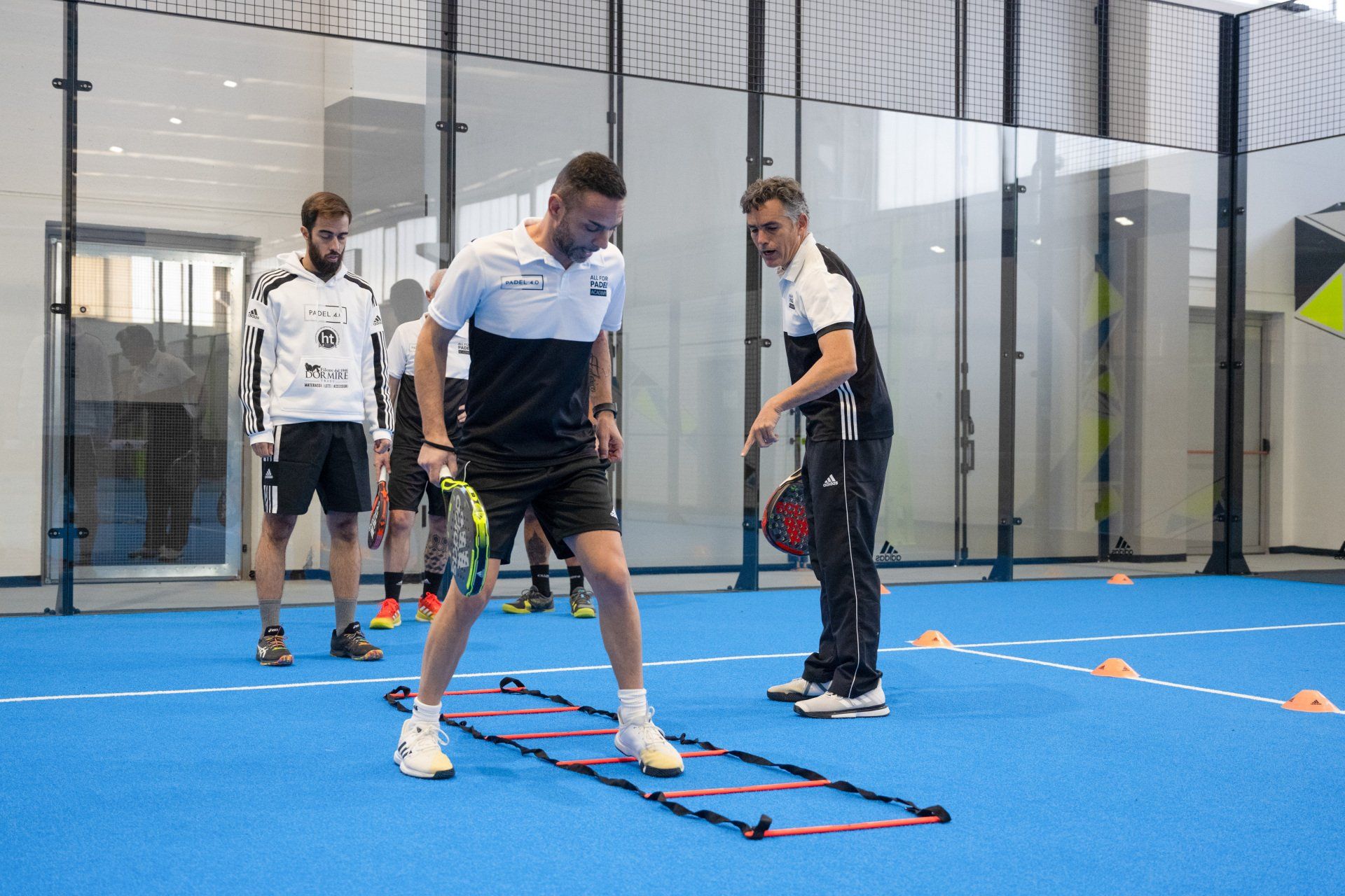 Un uomo in pantaloncini e polo si esercita con un allenatore su un campo blu, con altri giocatori sullo sfondo.