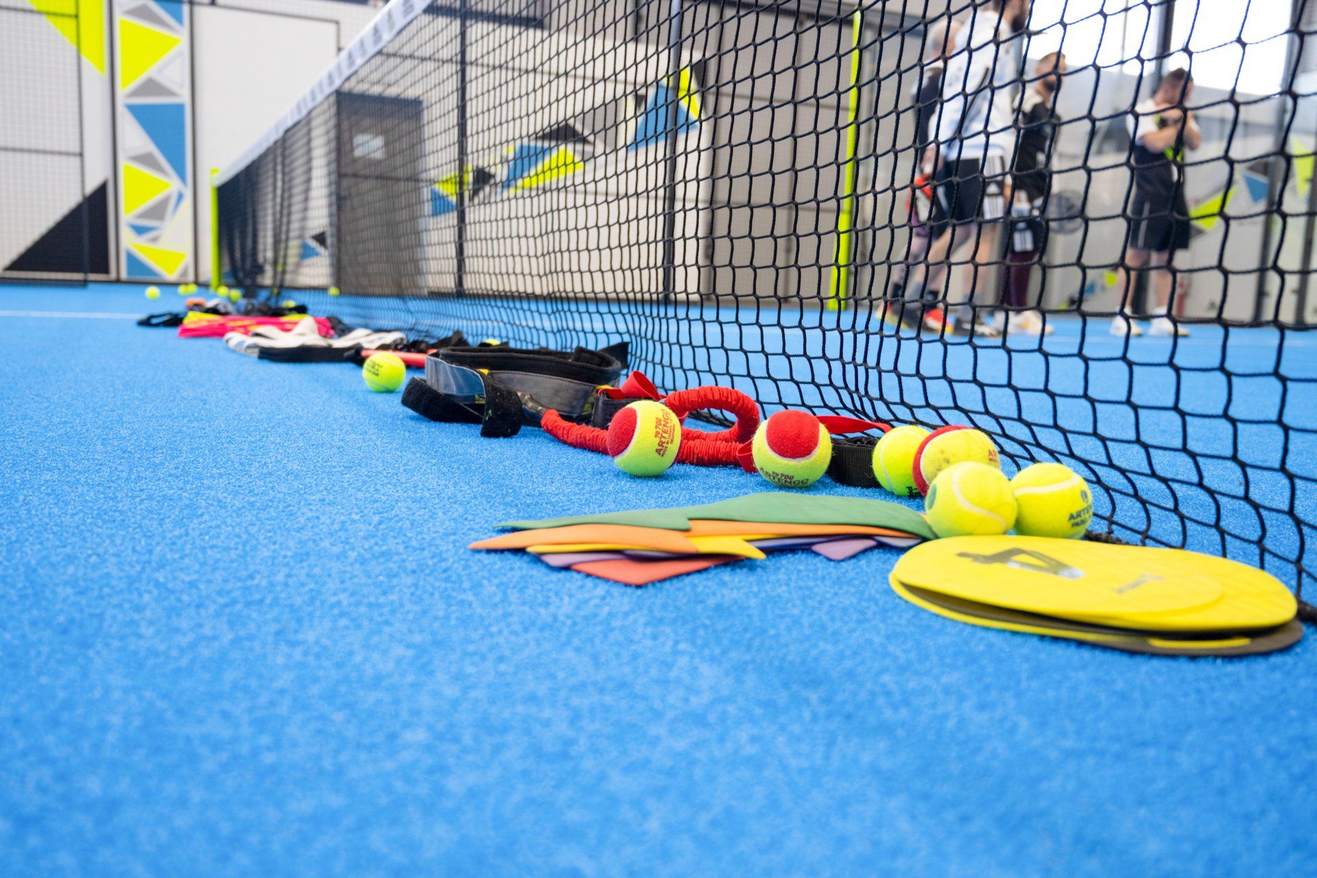 Campo da padel con attrezzatura: palline, racchette e rete. Persone in piedi sullo sfondo. Superficie del campo blu.