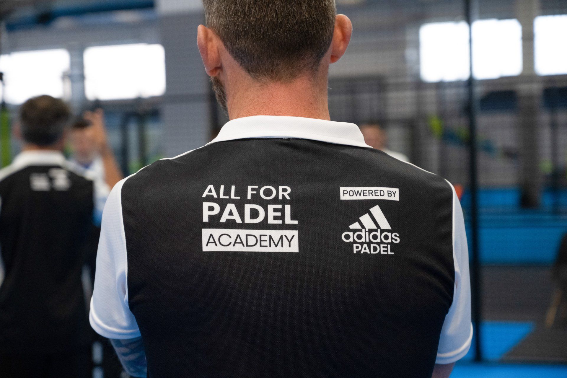 Schiena di una persona che indossa una maglietta nera Adidas Padel Academy in un campo da padel.