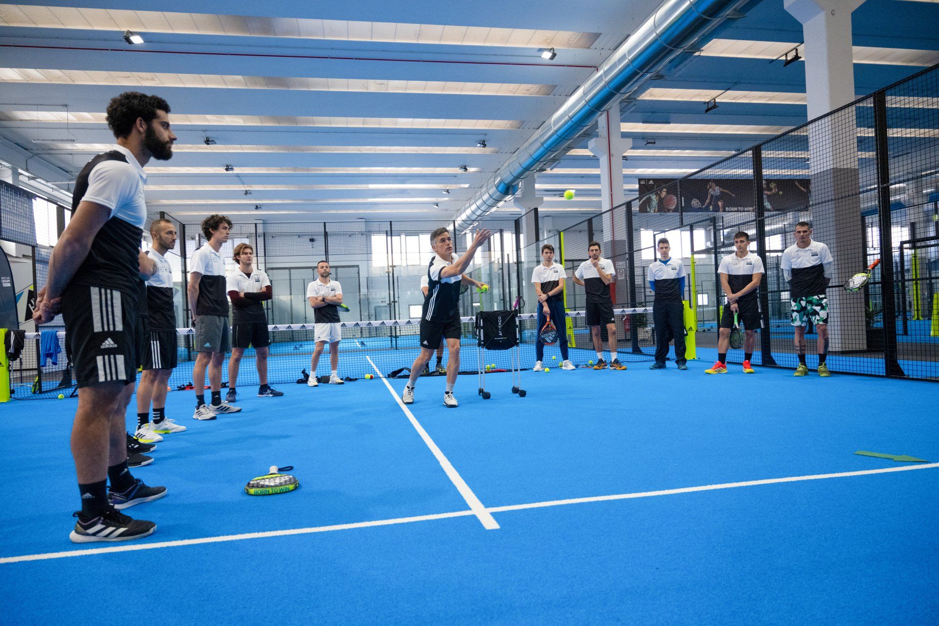 Sessione di allenamento di padel al coperto con l'istruttore che gesticola, i giocatori che osservano, campo blu.