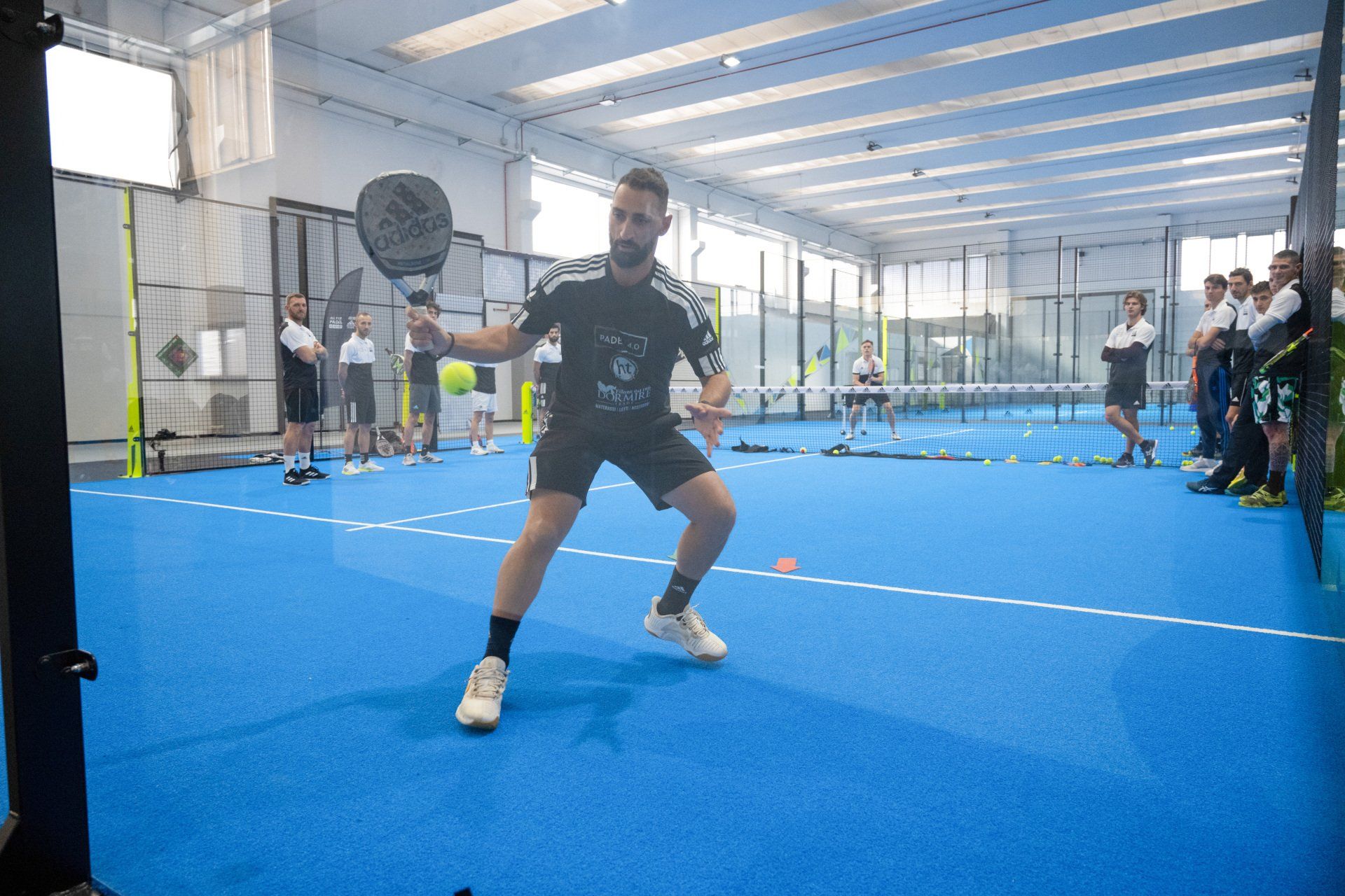 Un uomo in abbigliamento sportivo gioca a paddle tennis su un campo blu, mentre altri giocatori lo guardano sullo sfondo.