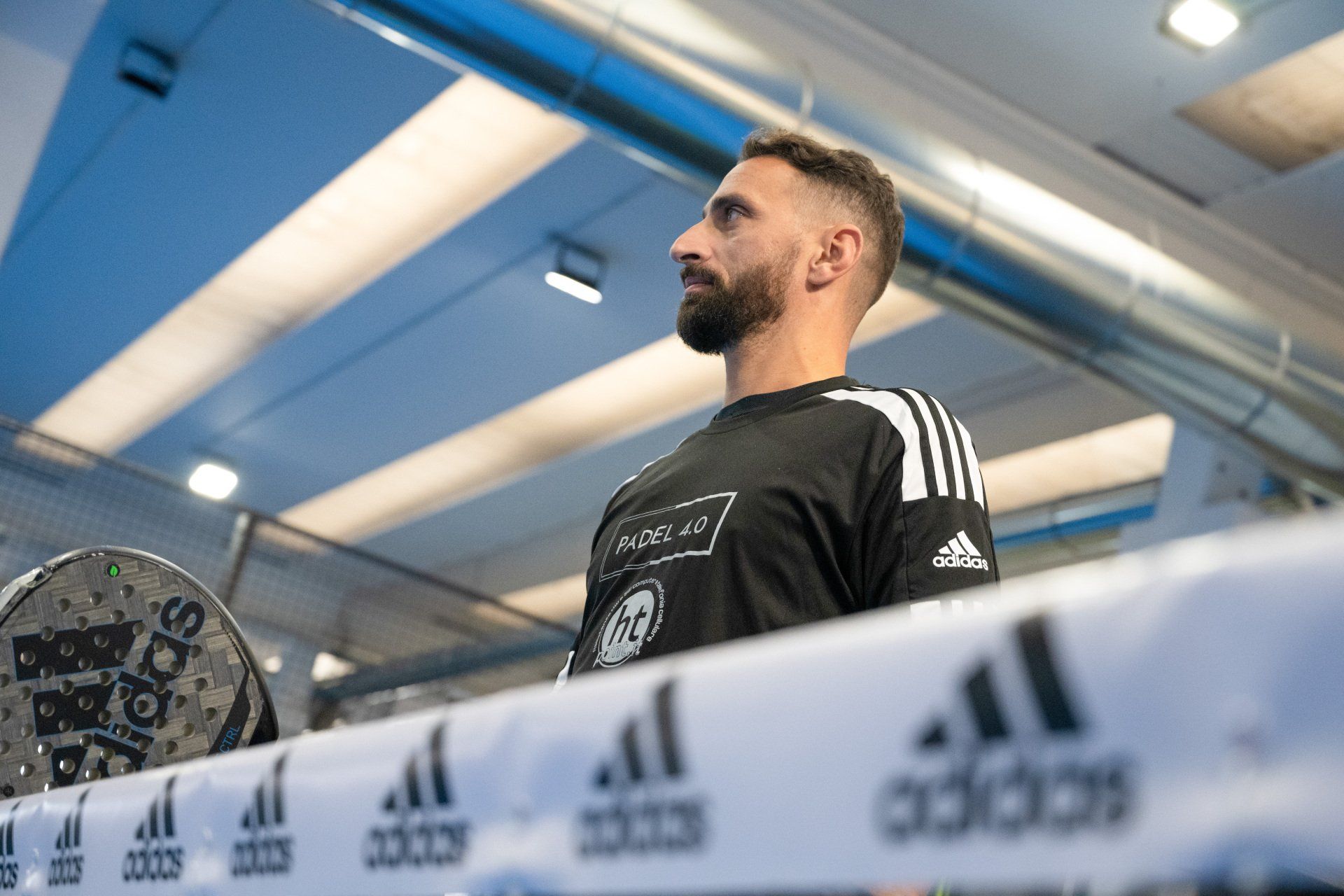 Un uomo con la barba e la maglietta nera guarda verso destra, vicino a uno striscione con il marchio Adidas in una palestra.