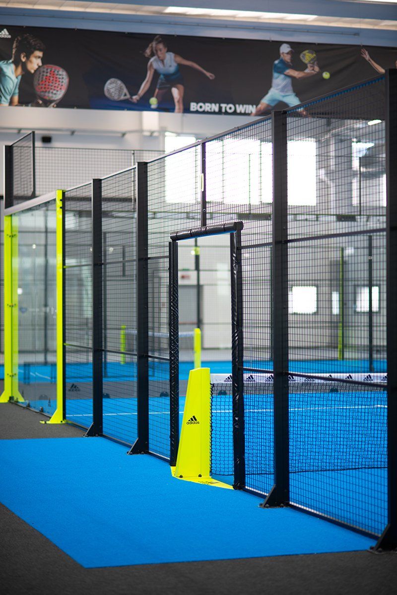 Campo da padel con superficie blu e barriere nere e gialle, in un ambiente al coperto.