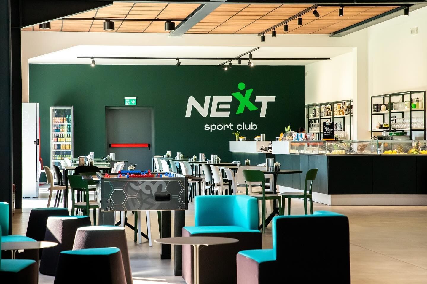 Interno del NEXT Sport Club: area salotto con tavoli, biliardino e bancone per il cibo in un ambiente moderno.