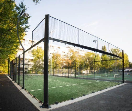 Campo da padel tennis all'aperto con pareti in vetro, erba verde e struttura in metallo nero sotto un cielo limpido.