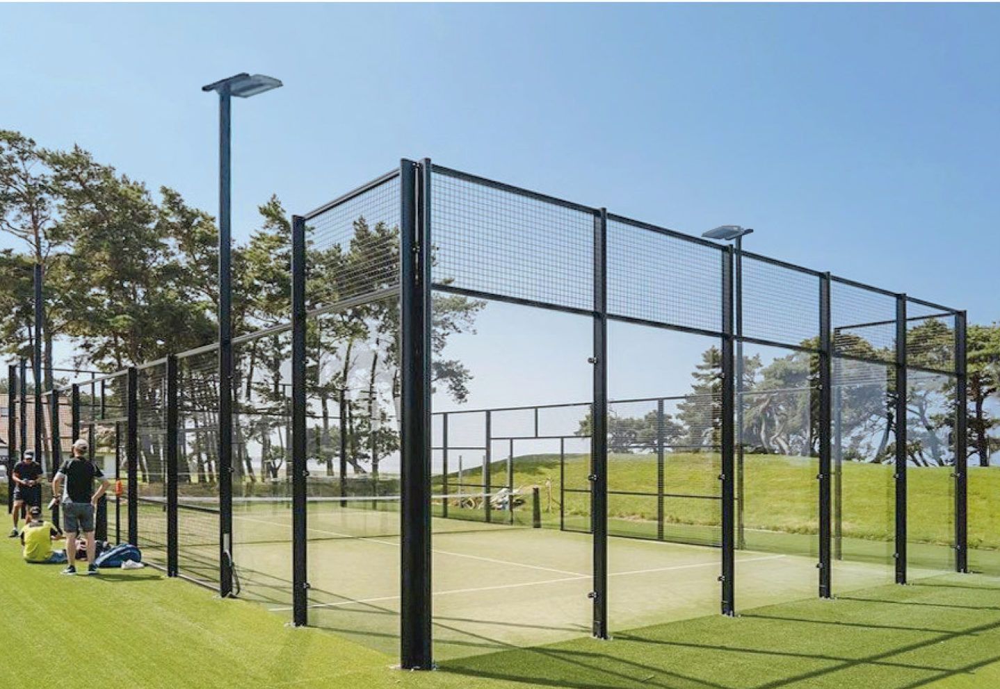 Campo da padel con giocatori, recinzione nera e illuminazione dall'alto in una giornata di sole.
