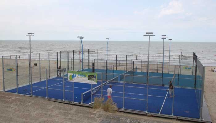 Campo da padel su una spiaggia con due giocatori, superficie blu e oceano sullo sfondo.