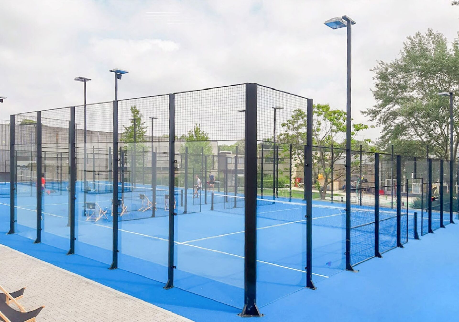 Campo da padel blu con recinzione nera, circondato da alberi e un cielo azzurro.