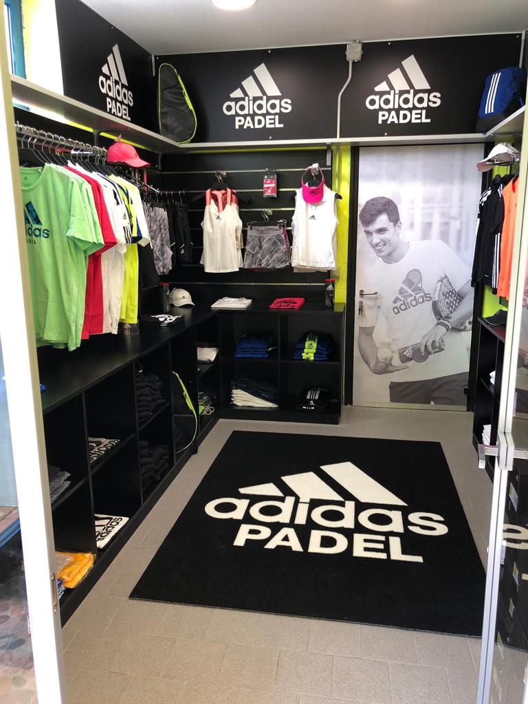 Interno del negozio Adidas Padel: scaffali neri, vestiti, scarpe e logo su tappeto e pareti.