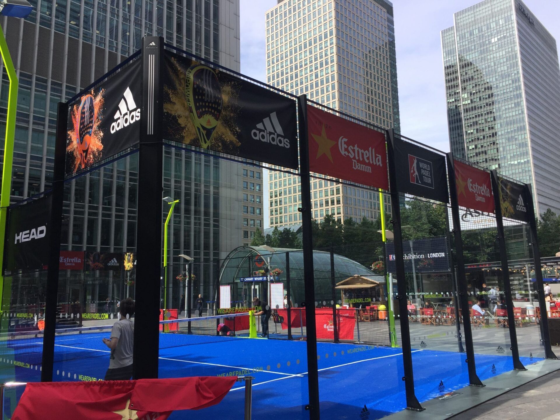 Campo da padel allestito all'aperto, sponsorizzato da Adidas ed Estrella Galicia. Campo blu, struttura nera, edifici alti.