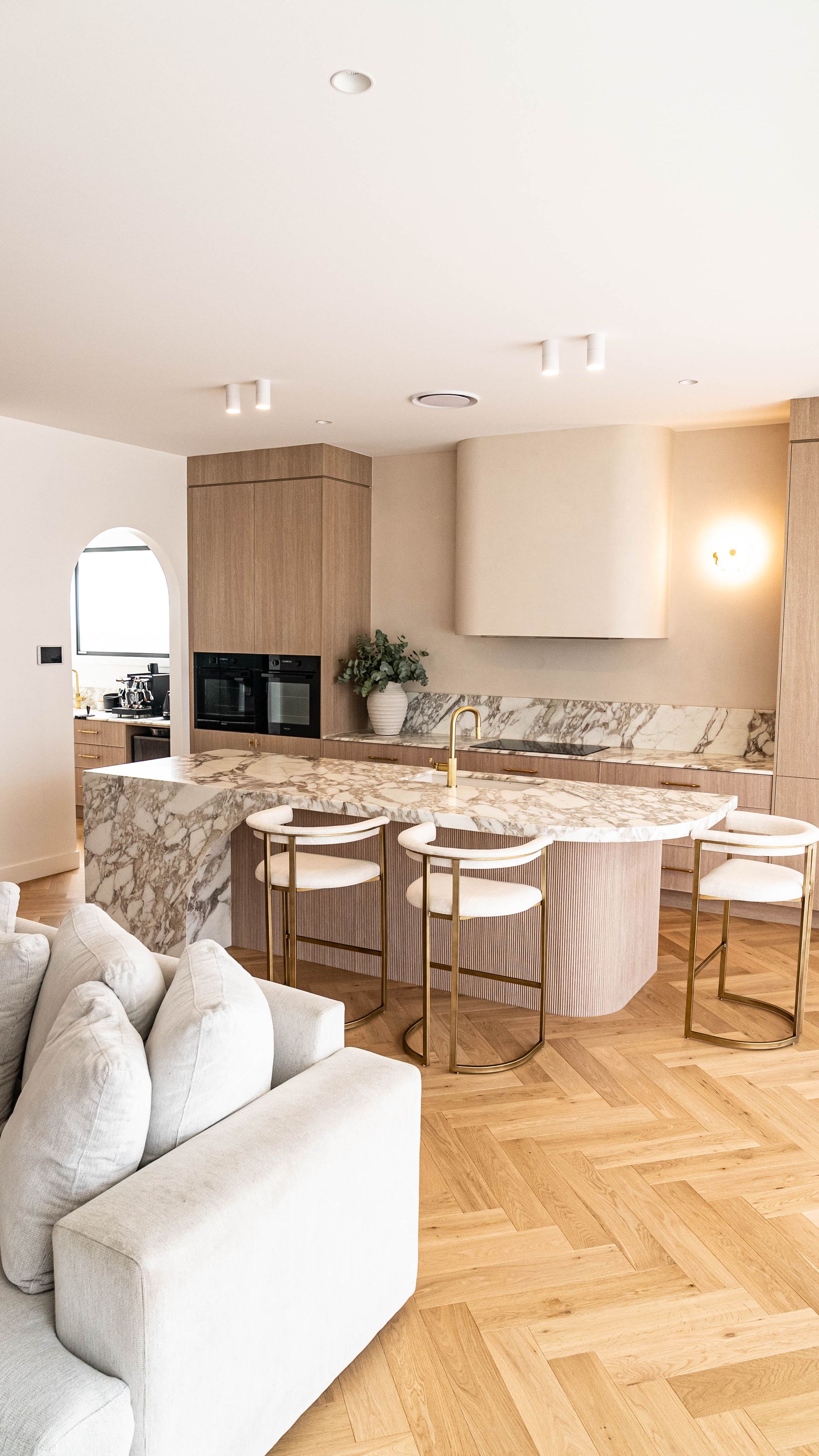 A modern kitchen with a large marble island, white bar stools, light wood cabinetry, and a herringbone wood floor.