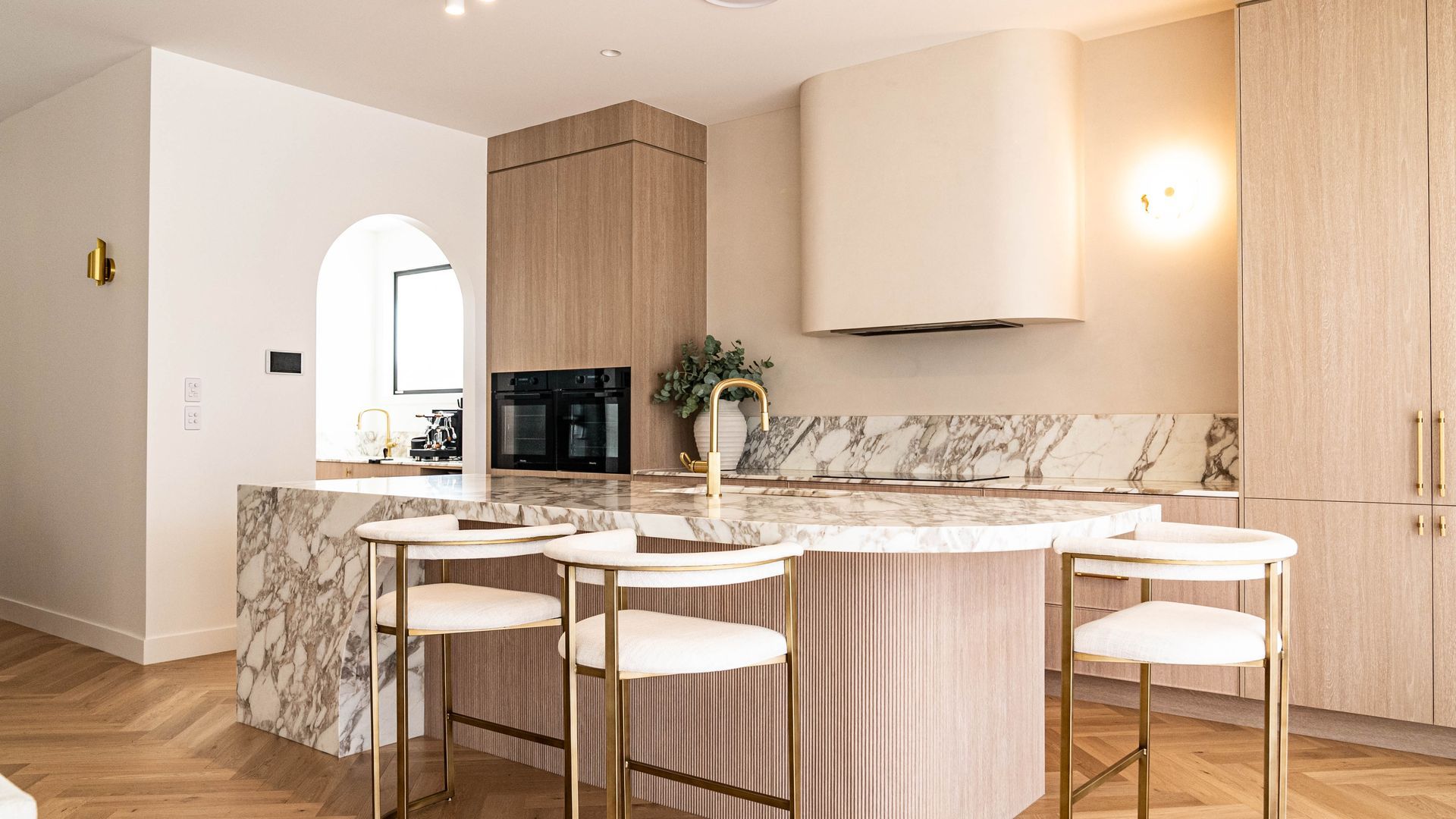 A bright, modern kitchen featuring a marble waterfall island with gold-accented white stools and light wood cabinetry.