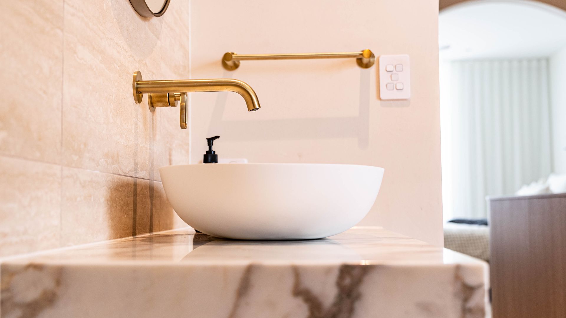 A white bowl sink sits on a marble countertop under a wall-mounted gold faucet in a modern bathroom.