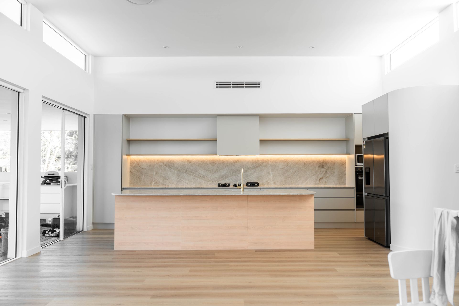 A minimalist kitchen with white walls, light wood flooring, a central wood-tone island, and marble backsplash with shelving.