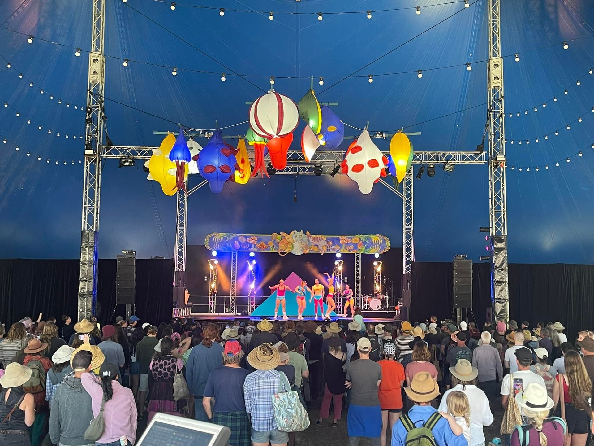 Stage performance at a festival: people watching under a blue tent. Colorful stage decor, performers on stage.
