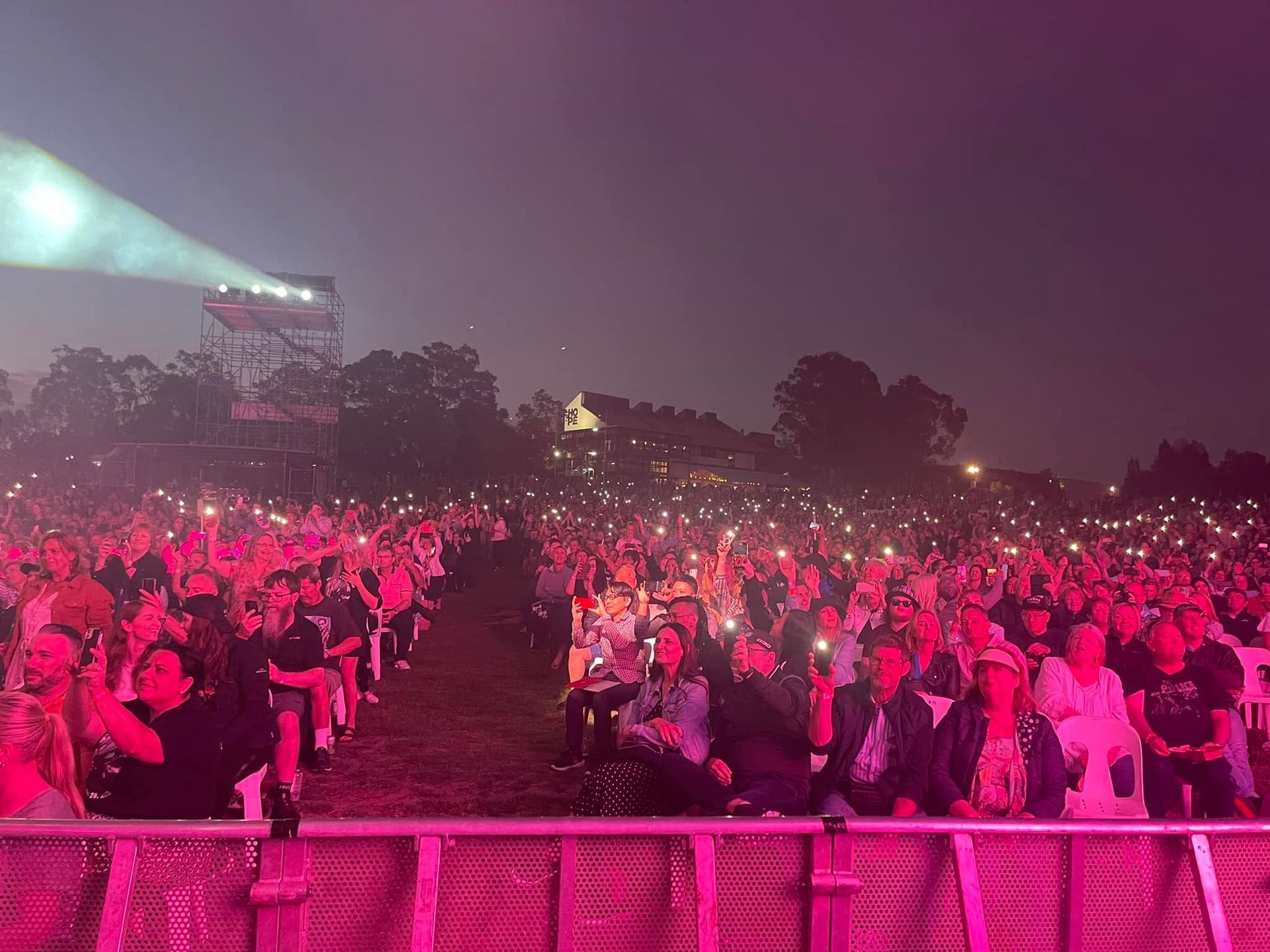 Large crowd at an outdoor concert, lit by pink lights and phone flashlights, stage in the background.