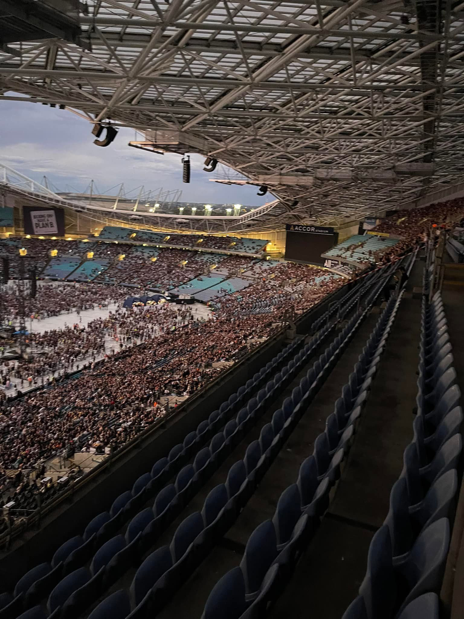 Large stadium filled with people, viewed from empty seats. Overcast sky above.