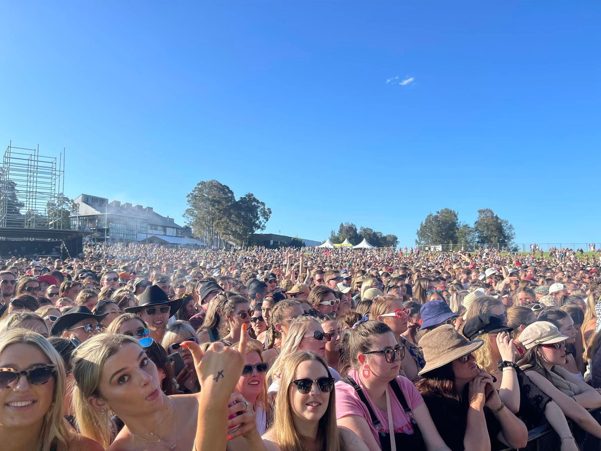 Large crowd at an outdoor concert, sunny day. Many people with sunglasses, some giving a 