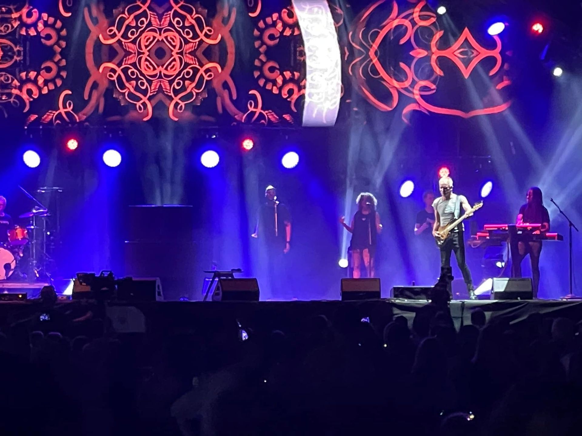 Band performing on stage with bright blue lights and a decorative screen behind them.