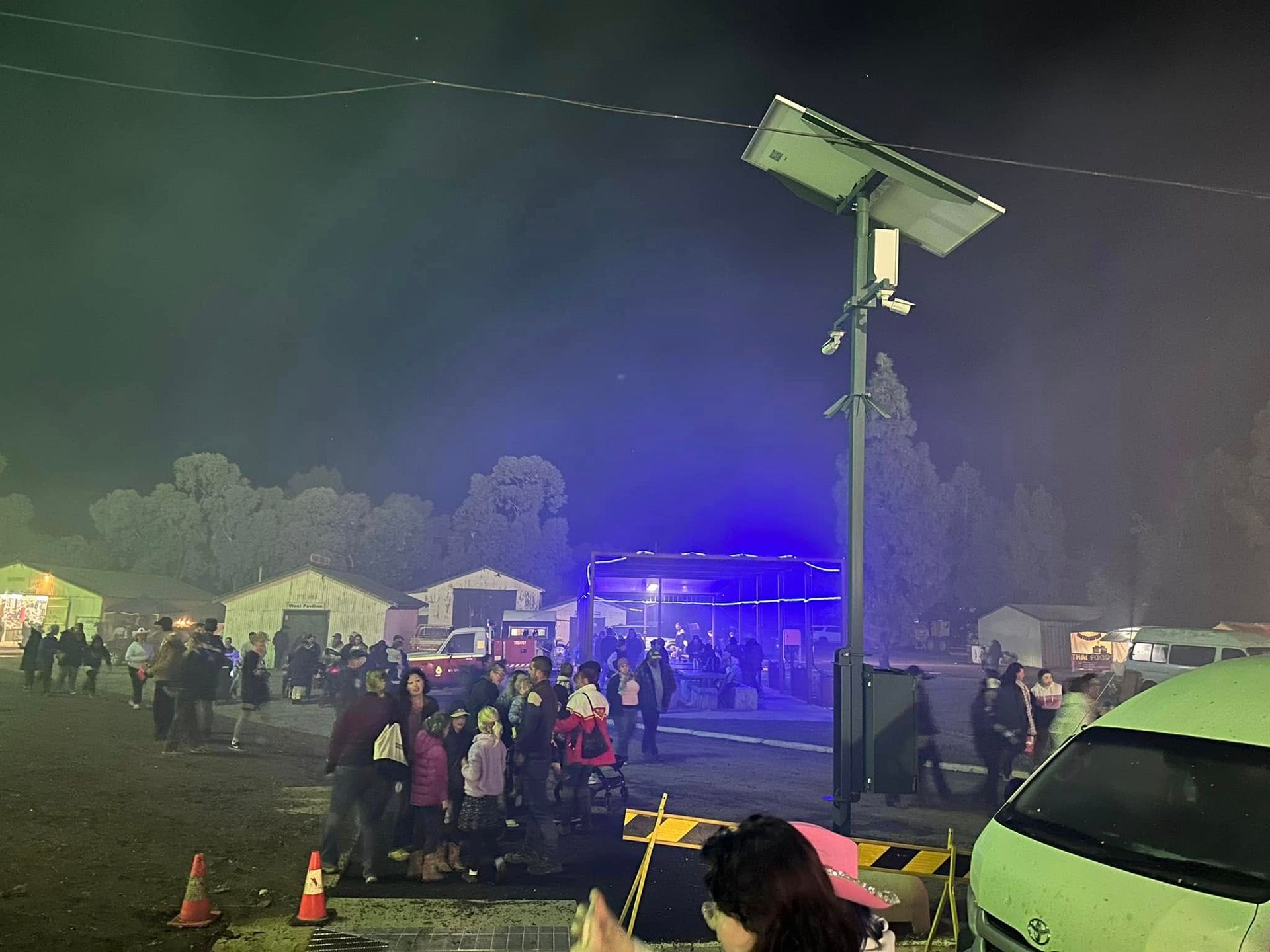 Night scene: Crowd outside festival tents with a security pole, lights, and vehicles.