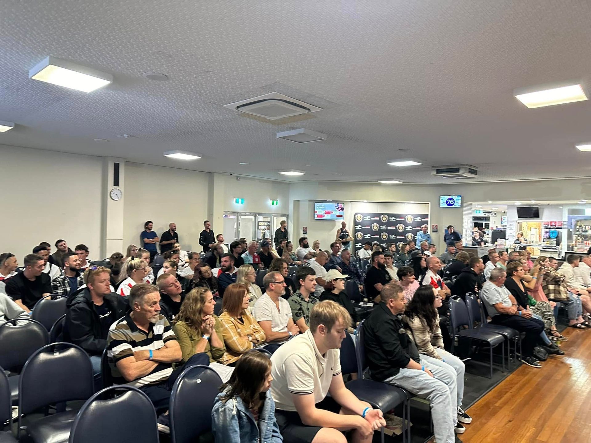 Audience in a large room, listening intently. Many people sitting in rows of chairs.