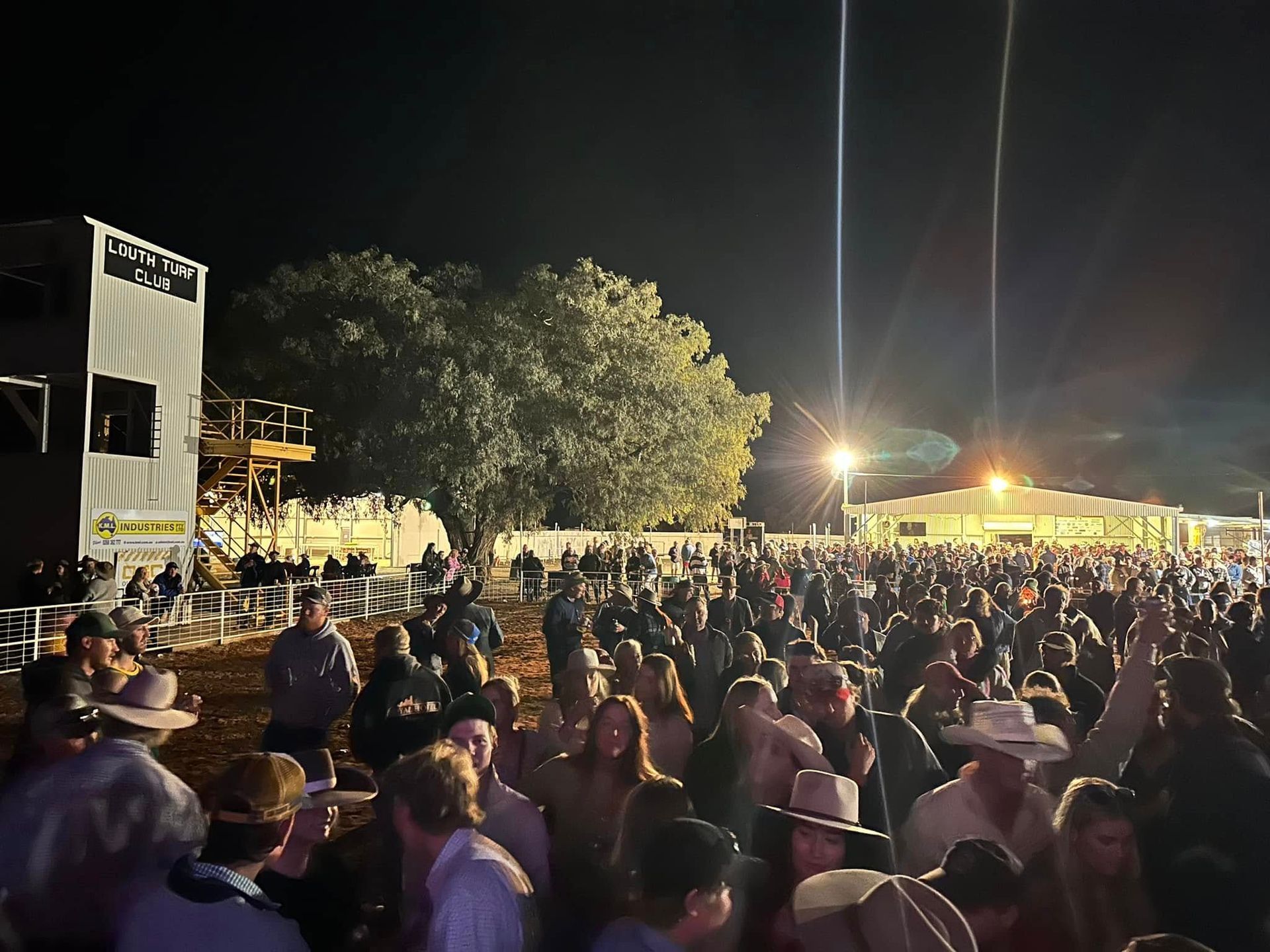 Large crowd at a nighttime outdoor event with a stage and lights, many wearing cowboy hats.