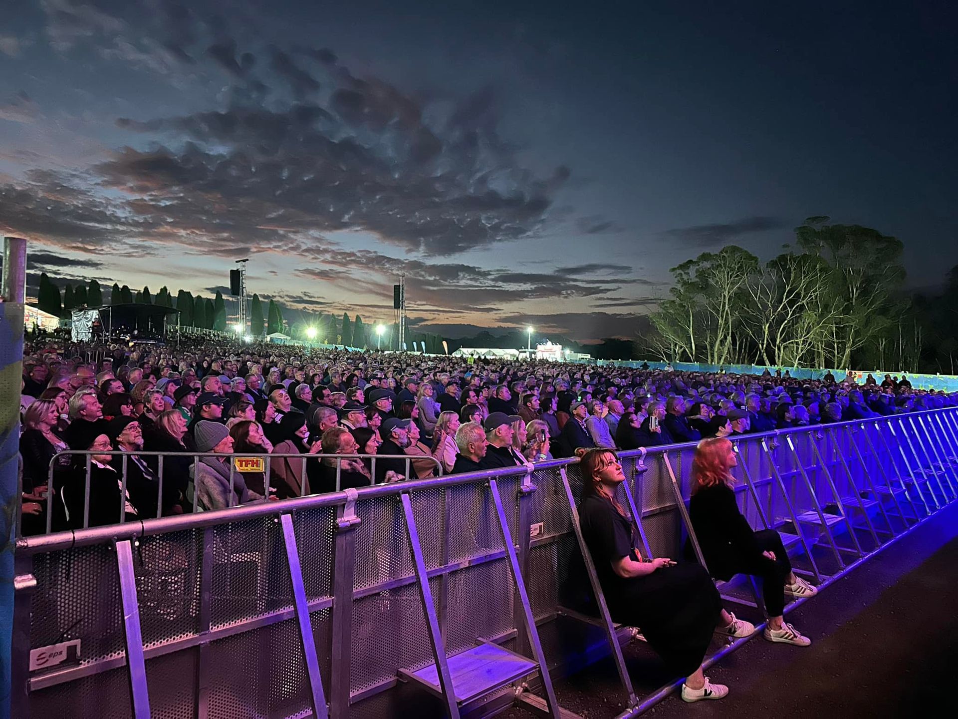 Large crowd at an outdoor concert, watching the stage. Evening sky with clouds. Metal barricade in foreground.
