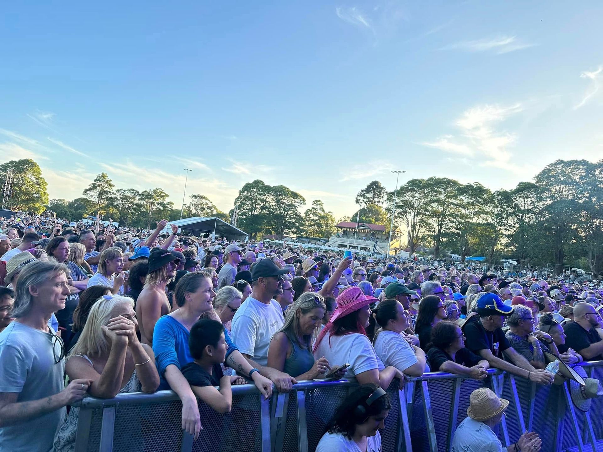 Crowd at an outdoor concert, watching the stage. People standing close, some wearing hats, under a blue sky.