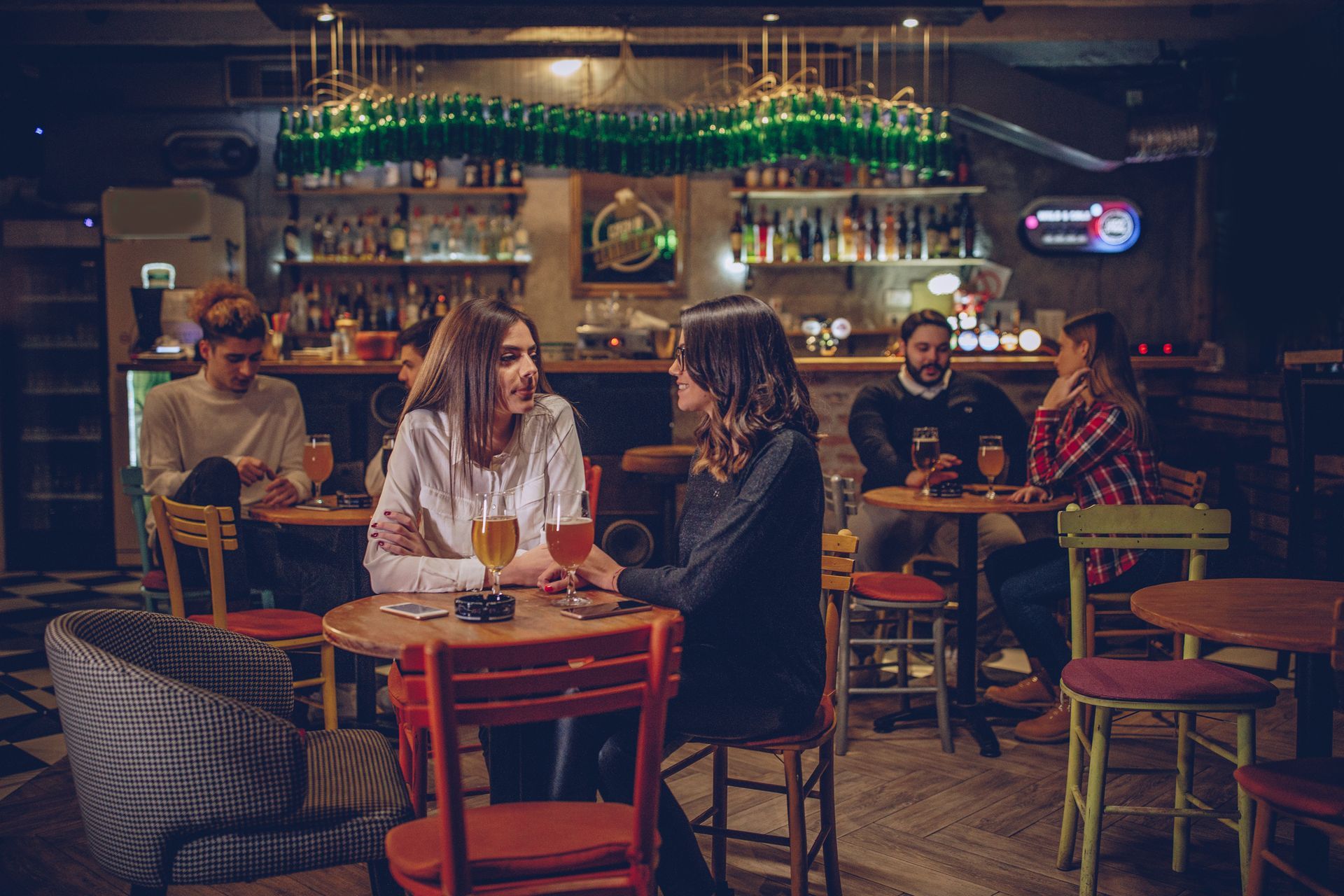 People at a bar: two women talking, others drinking, relaxed atmosphere with dim lighting.