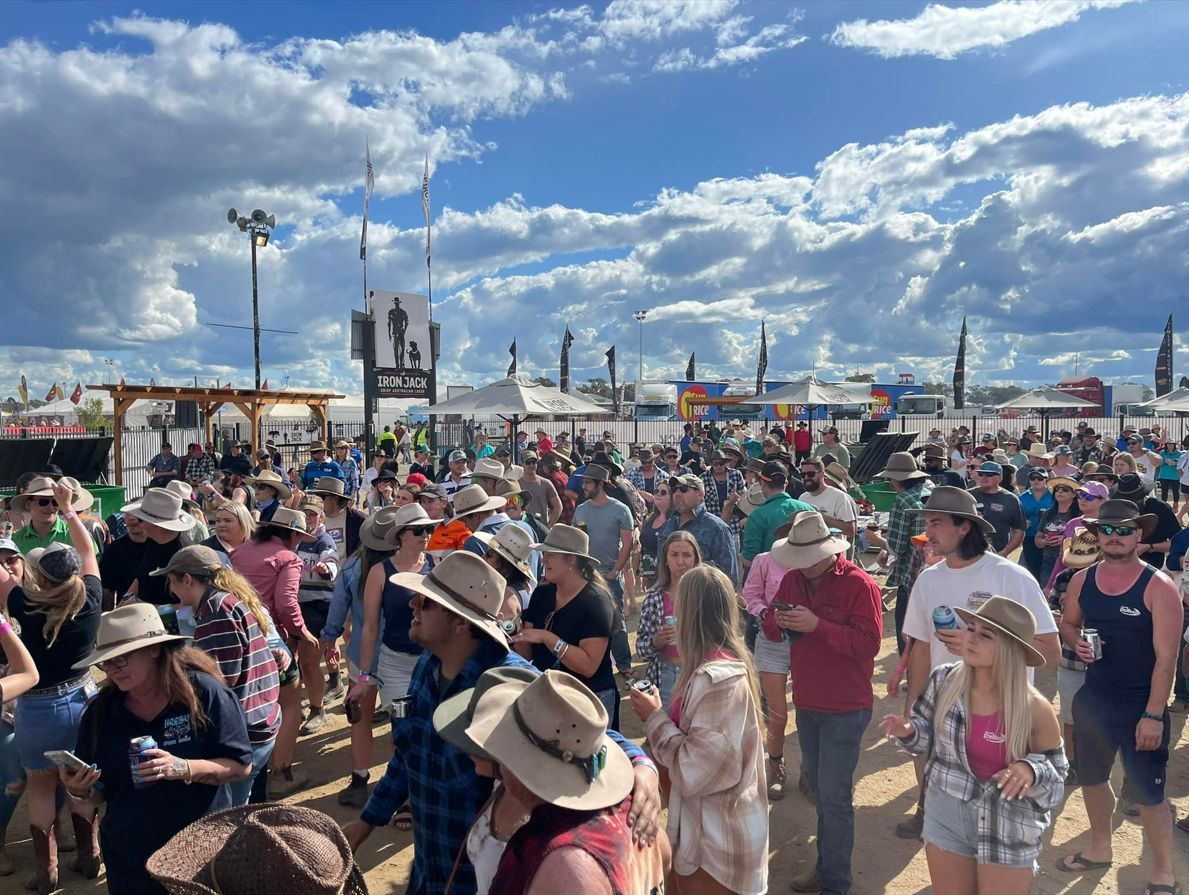 Large crowd at an outdoor festival. People wearing hats, mingling under a cloudy blue sky.