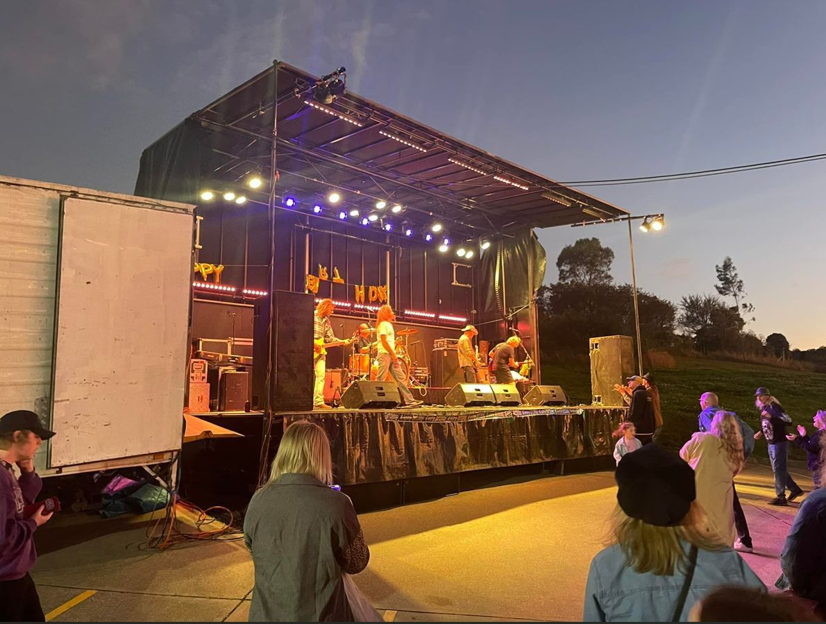 Stage with band performing outdoors at dusk; people in the audience. Stage lit with colorful lights.