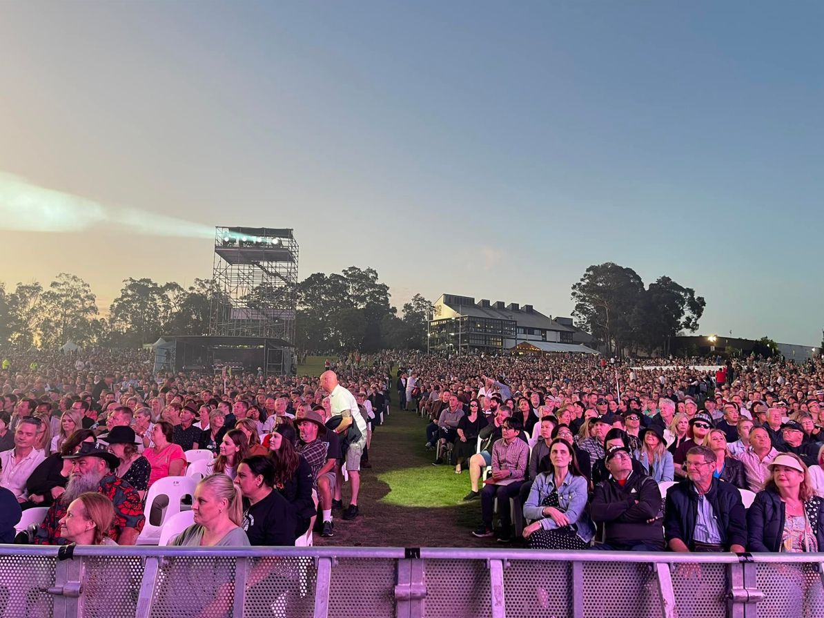 Large crowd at outdoor concert; stage in the distance.