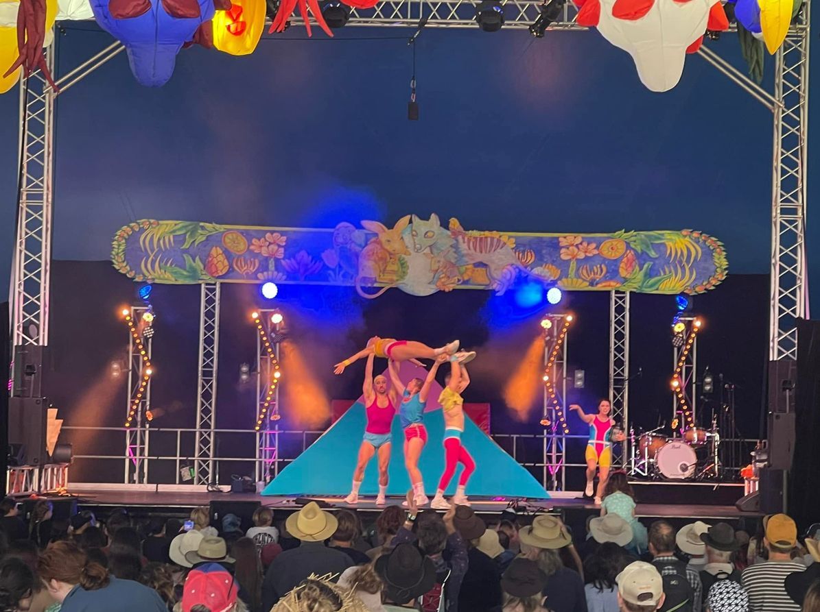Acrobats perform on stage under colorful lights. Three acrobats hold another person aloft. Audience in foreground.