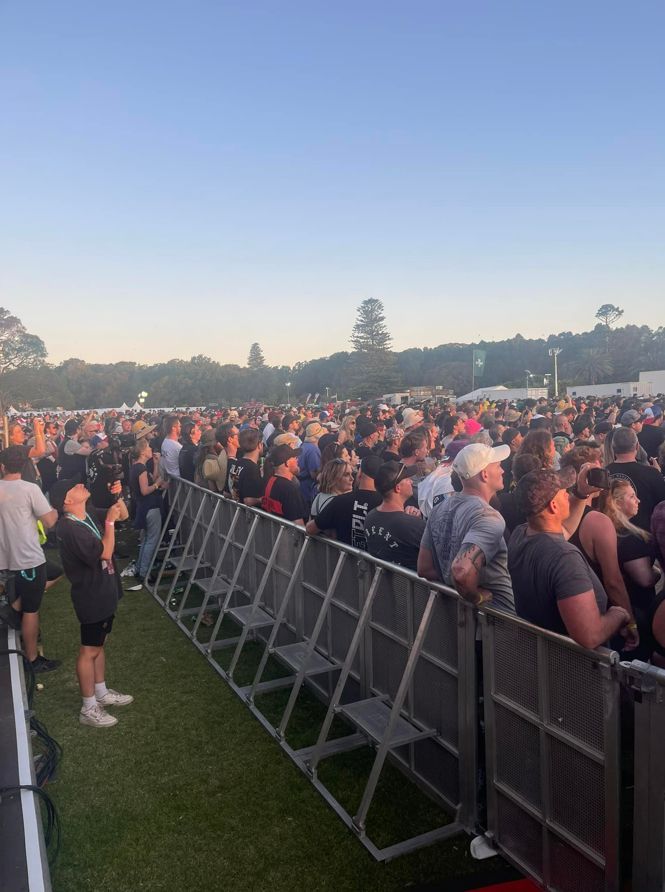 Crowd at an outdoor concert, near metal barricades. People are watching the stage, under a clear sky.