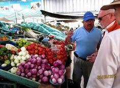 Two men are standing in front of a vegetable stand at a market.