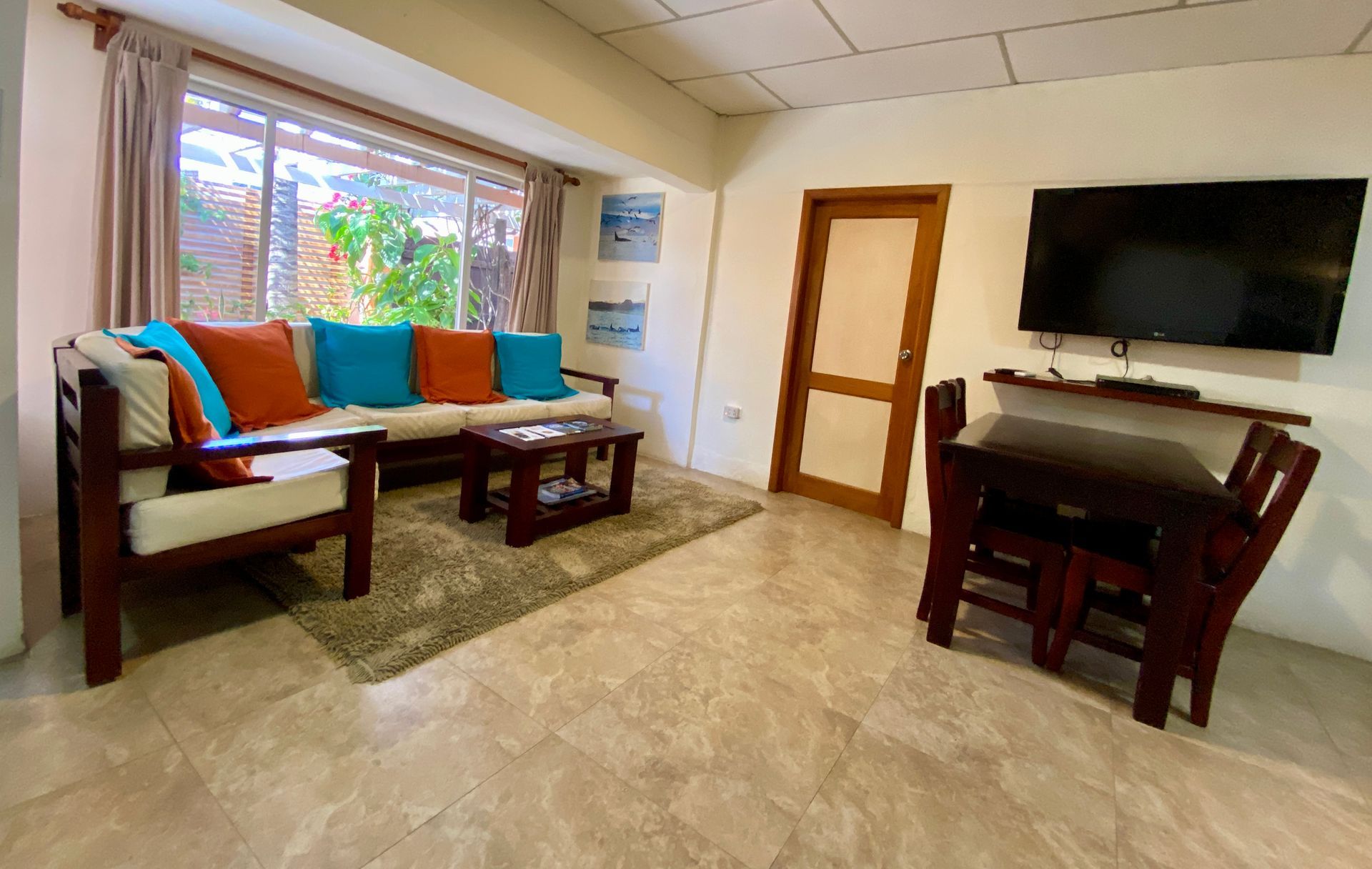 Living room with a sofa, coffee table, dining set, and TV. The room has tan walls and flooring and a large window.