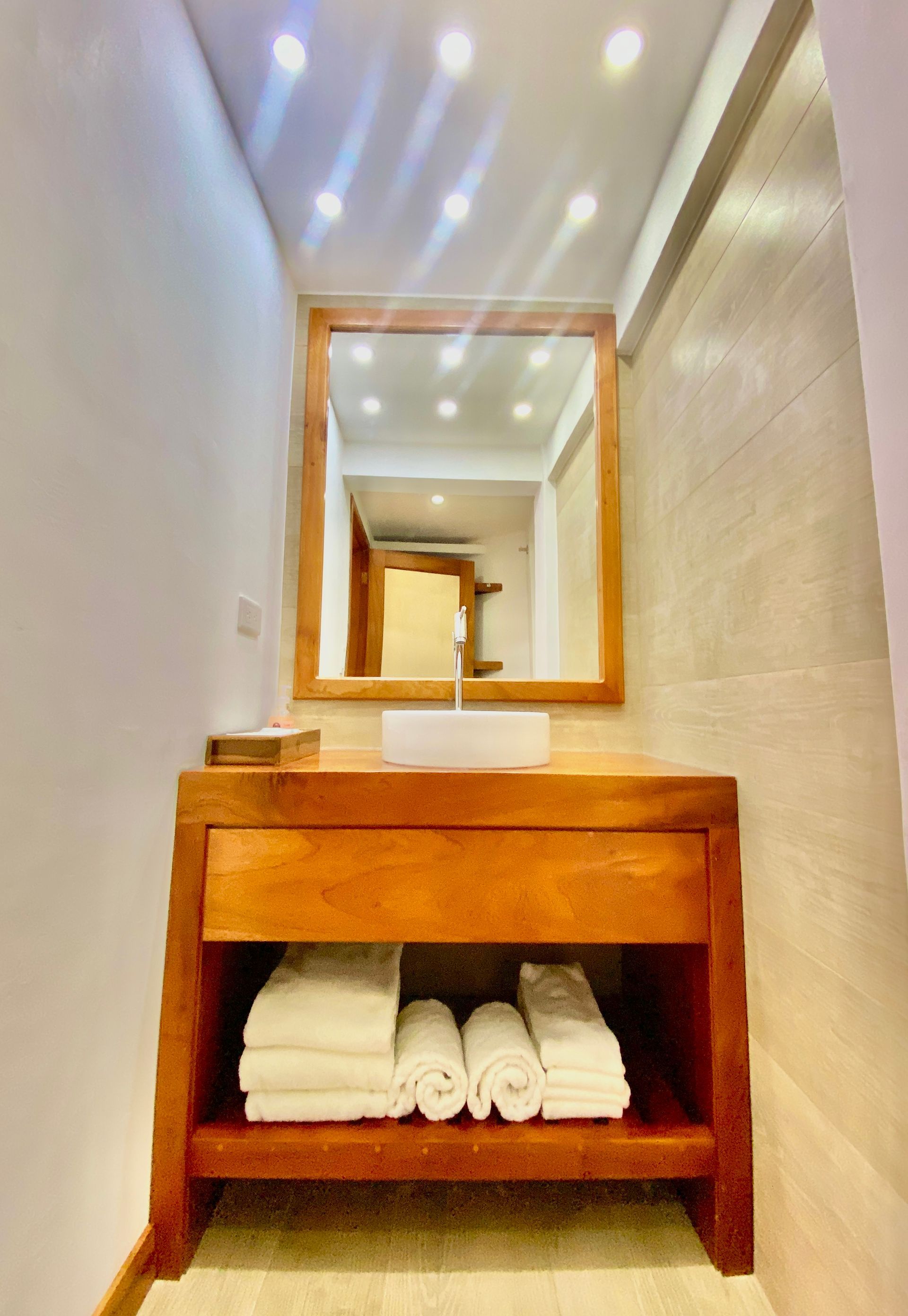 A wooden vanity with a mirror, a white sink, and rolled white towels sits in a narrow bathroom.